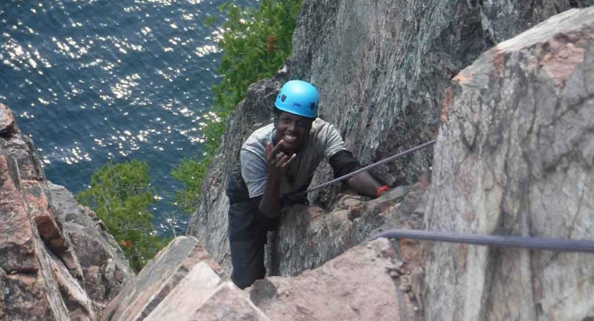 an outward bound student who is rock climbing above a body of water smiles at the camera